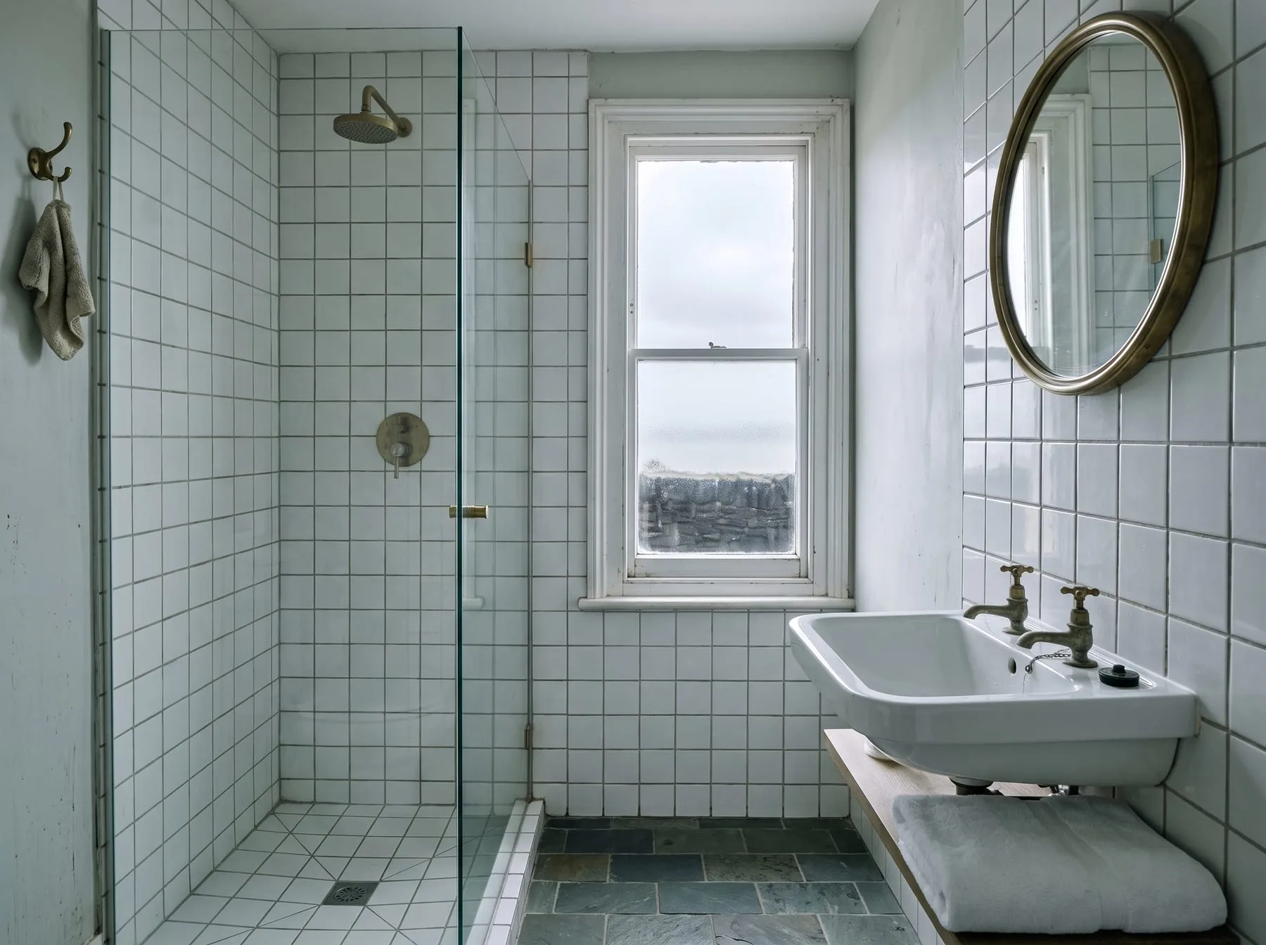 The bathroom, with a walk-in shower behind glass, white square tiles and a small window