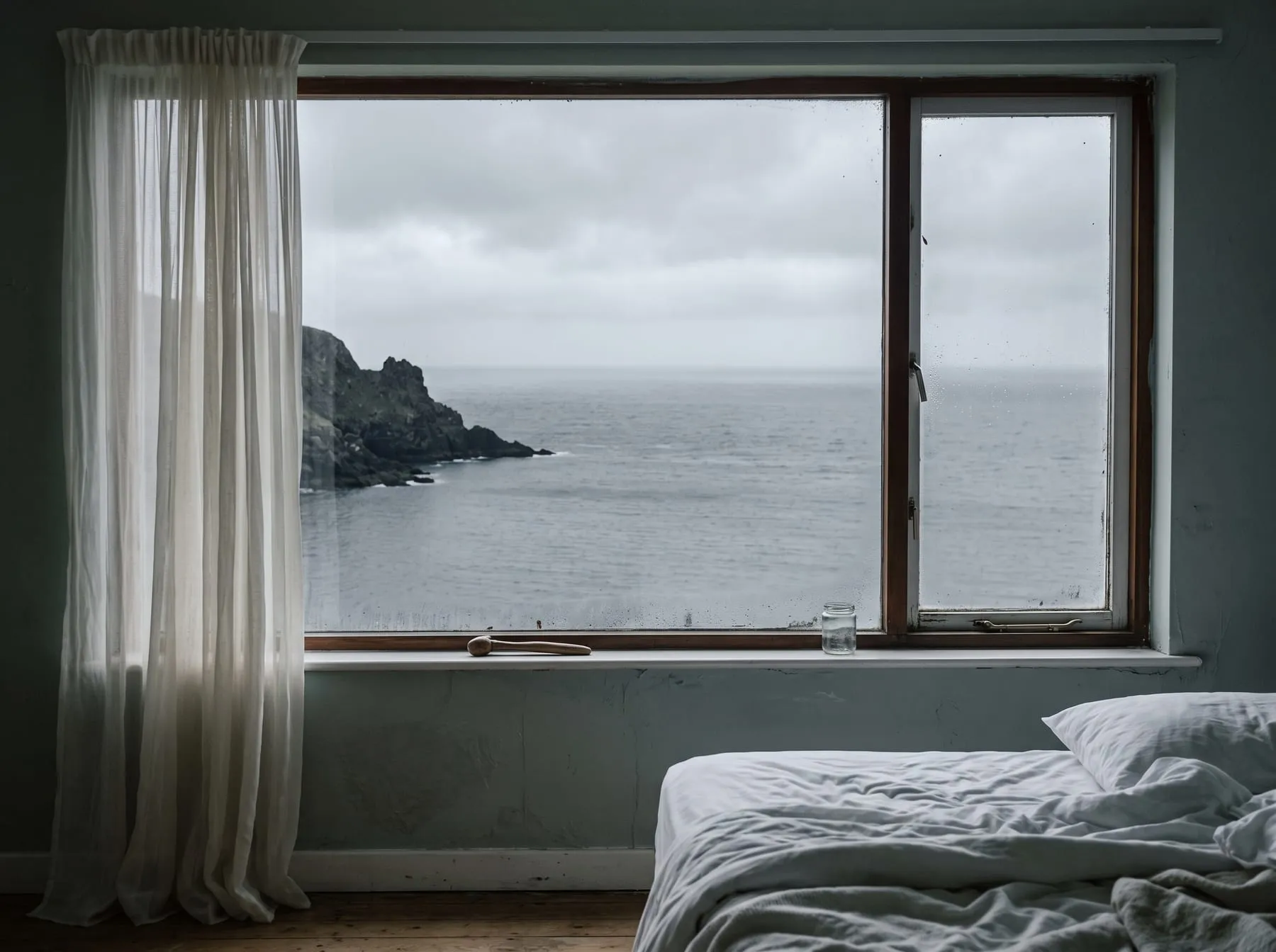 The view north from the bed: flat gray sea, a dark headland, and the curtain drawn to one side